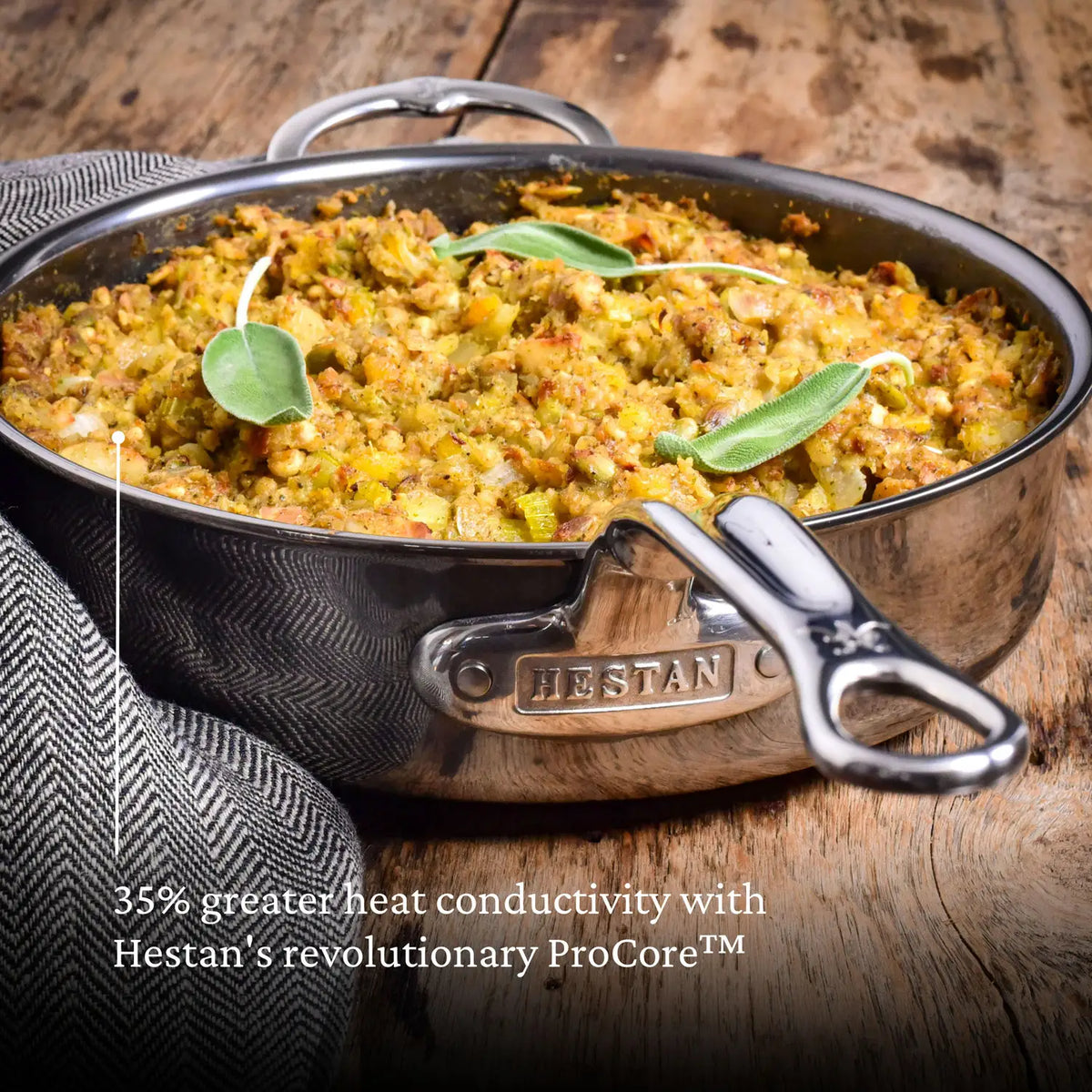 A Titanium Ultimate Cookware dish by NanoBond, filled with casserole and herbs, rests on wood. Its handle features the brand logo. 35% greater heat conductivity with Hestans revolutionary ProCore™ is highlighted in a text overlay.