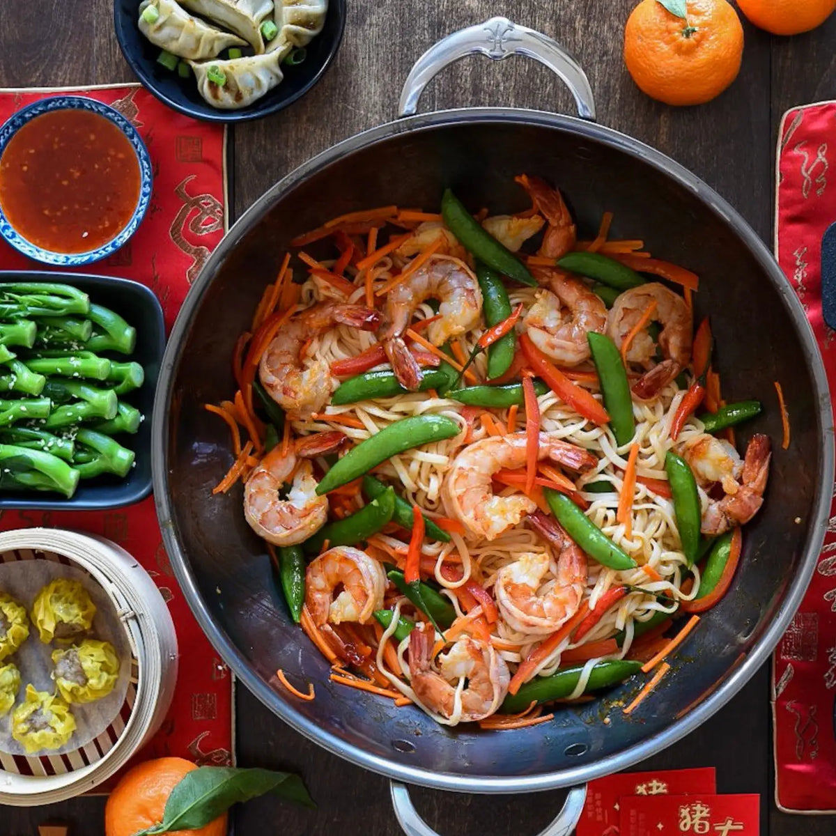 A bowl of stir-fried noodles with shrimp, snap peas, and carrots is on a table. Nearby are plates of dumplings, steamed greens, two tangerines, and a bamboo steamer with dim sum. The setting is completed by a NanoBond 14-Inch Titanium Wok for high-heat cooking on red mats.