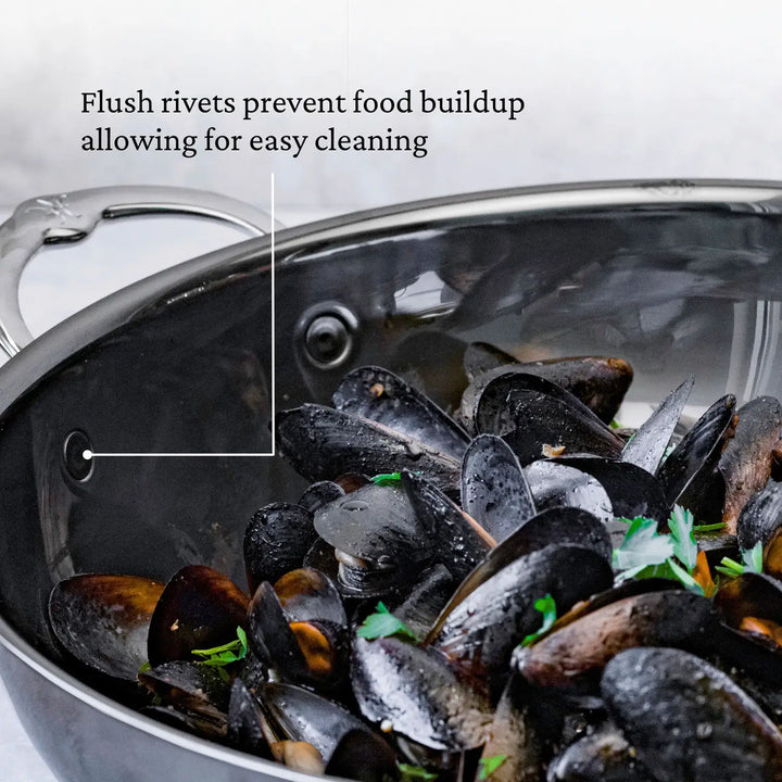 The 14-Inch Titanium Wok by NanoBond, with its sleek molecular titanium finish, showcases exceptional cooking performance and high-heat tolerance. Designed with flush rivets and labeled arrows to prevent food buildup, this stainless steel wok is durable and perfect for cooking mussels garnished with parsley.