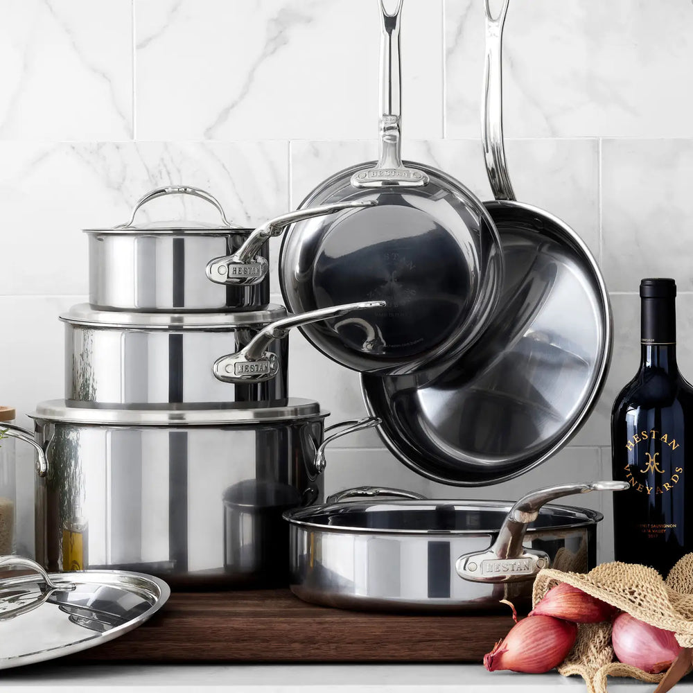 A 10-Piece Titanium Ultimate Cookware Set from NanoBond®—featuring pots, pans, and lids crafted with molecular titanium for exceptional durability—is displayed on a wooden surface. Beside this innovative set are a bottle, shallots, and garlic, all arranged in front of a tiled backsplash.