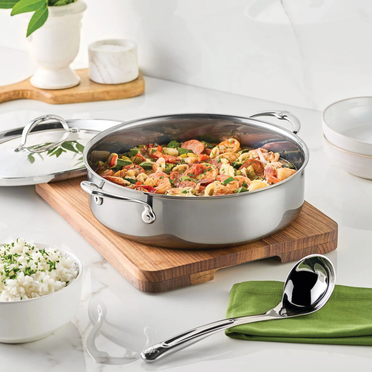 A NanoBond Titanium Rondeau with lid holds a cooked mixed dish on a wooden cutting board beside a bowl of white rice, with a metal serving spoon on a green napkin and kitchen items arranged on a white countertop.