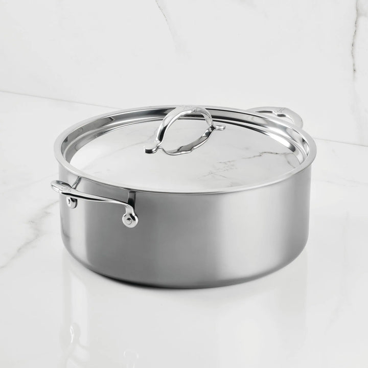 A NanoBond Titanium Rondeau with Lid sits on a white marble countertop. Its metal handles and reflective finish highlight the elegant molecular titanium cookware design against the marble wall.