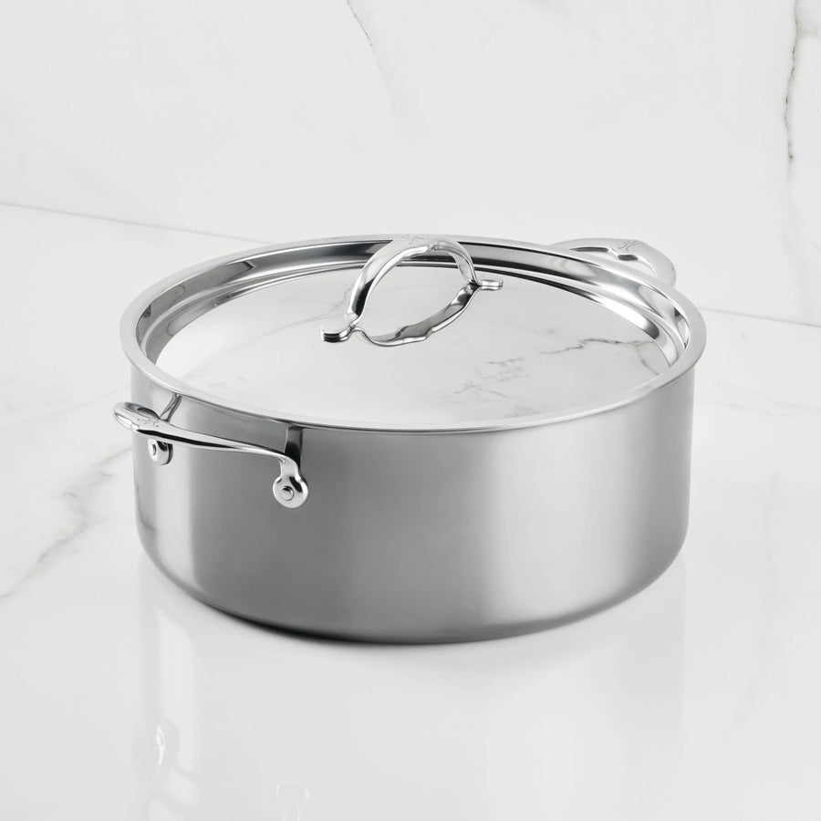 A NanoBond Titanium Rondeau with Lid sits on a white marble countertop. Its metal handles and reflective finish highlight the elegant molecular titanium cookware design against the marble wall.