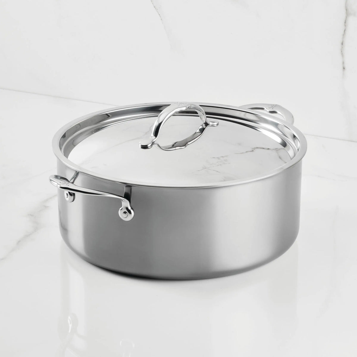 A NanoBond Titanium Rondeau with Lid sits on a white marble countertop. Its metal handles and reflective finish highlight the elegant molecular titanium cookware design against the marble wall.
