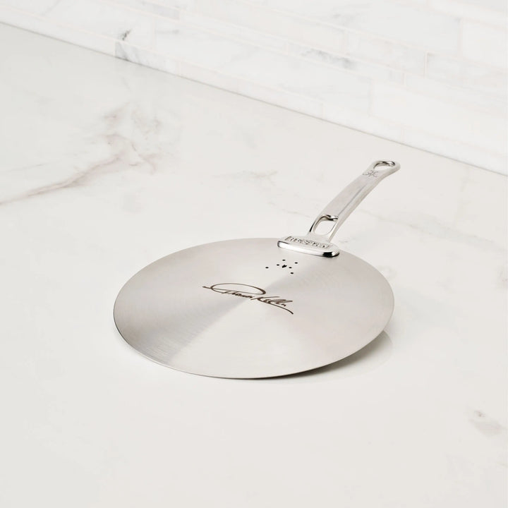 The Thomas Keller Commercial Clad Stainless Steel Universal Lid, featuring an engraved handle, sits on a white marble countertop—an ideal addition to your eco-friendly cookware collection.