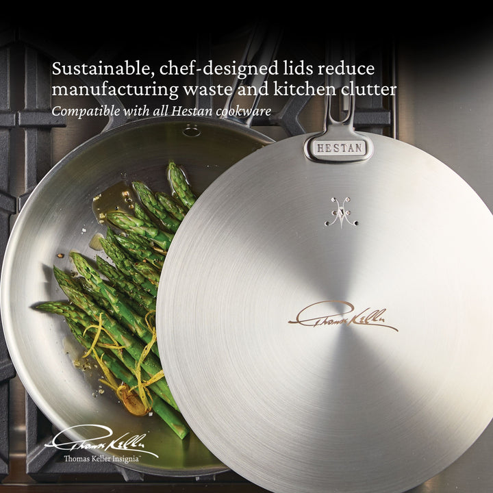 A Thomas Keller Commercial Clad Stainless Steel Universal Lid, featuring his signature, sits next to a stainless steel pan with asparagus and herbs. Sustainable and chef-designed, the lids fit seamlessly with Hestan cookware.