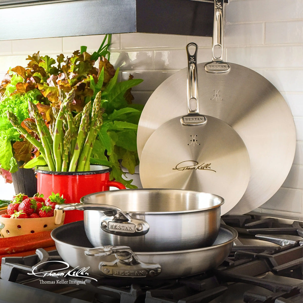 Two Thomas Keller Commercial Clad pans and two Stainless Steel Universal Lids are stacked on a stove. Behind them, a red pot holds asparagus and greens beside a bowl of strawberries, with kitchen utensils and a white tiled wall in view.
