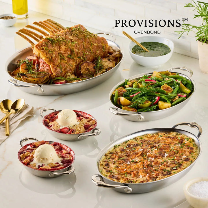 A table features a roasted rack of meat, veggies, berry cobblers with ice cream, and green sauce. The baked casserole is served in the Hestan Provisions 6.5" x 5.5" Oval Au Gratin Set (2-piece).