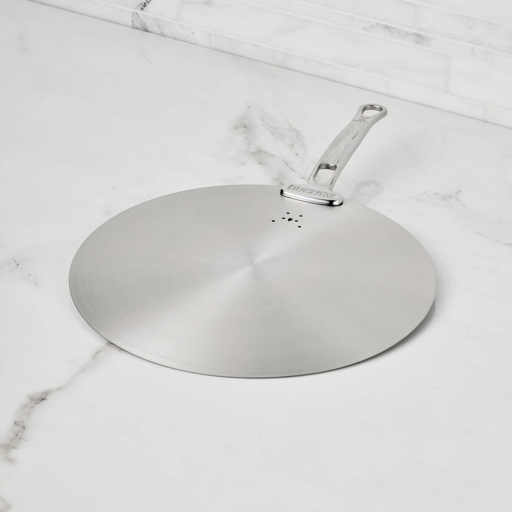 The Hestan Provisions Stainless Steel Universal Lid serves as both a splatter screen and lid, features a handle, rests on a white marble countertop with gray veining, is eco-friendly, and dishwasher safe—an essential for any kitchen.