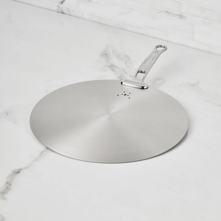 The Hestan Provisions Stainless Steel Universal Lid serves as both a splatter screen and lid, features a handle, rests on a white marble countertop with gray veining, is eco-friendly, and dishwasher safe—an essential for any kitchen.