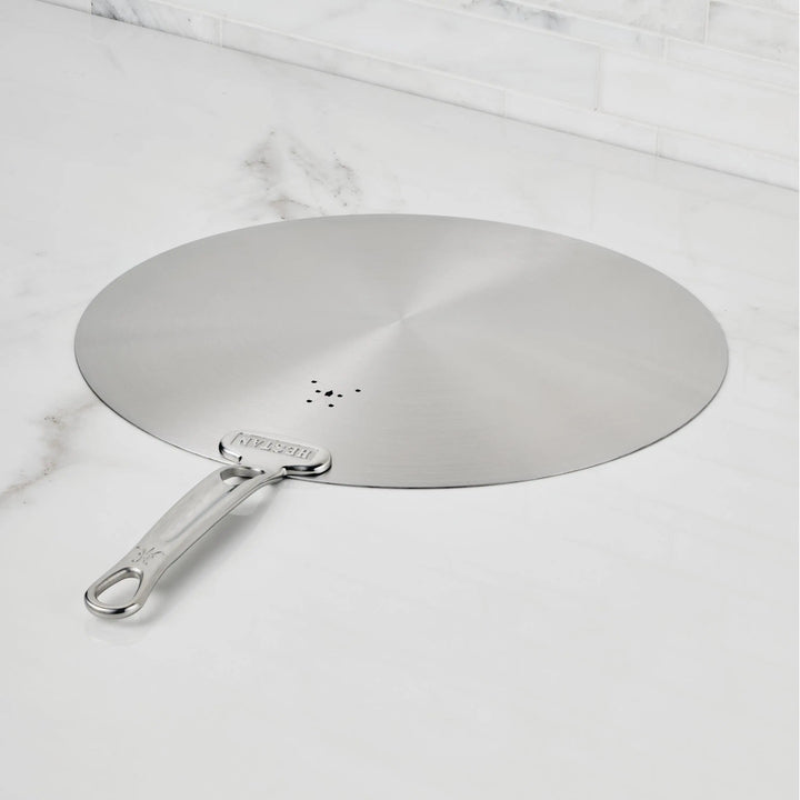 The Hestan Provisions Stainless Steel Universal Lids feature a long handle, central small holes, and can serve as a splatter screen—an eco-friendly, dishwasher safe kitchen essential displayed on a white marble countertop.