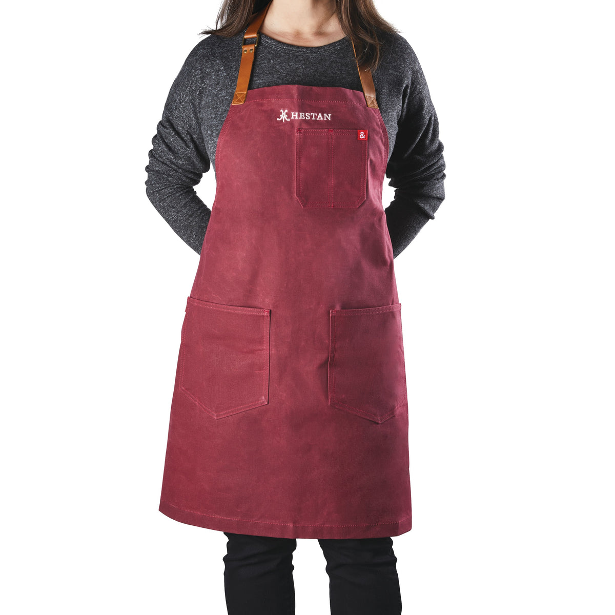 The person is wearing a dark sweater and the red Hestan x Hedley & Bennett Grilling Apron with pockets and elegant leather neck straps. Crafted from waxed canvas by Accessories, it features a logo near the chest, as they stand against a white background.