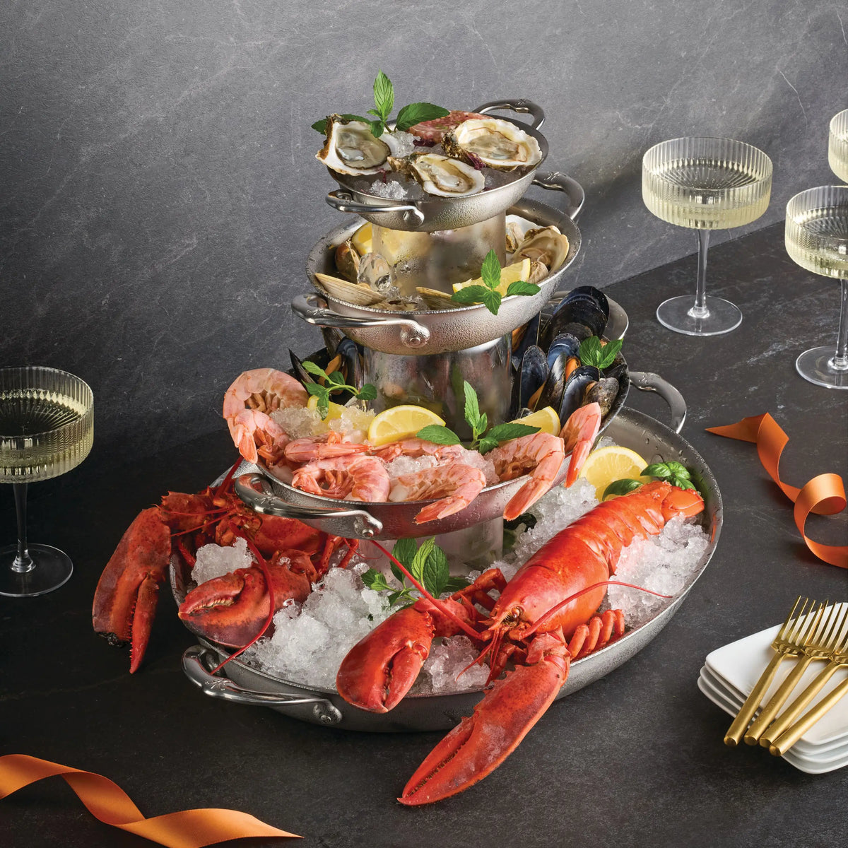 A three-tiered seafood platter on ice is served with lemon wedges and herbs, next to three pale drinks, gold forks, and the Hestan Provisions 17.5-Inch x 14-Inch Clad Oval Roaster, all presented on a dark surface.