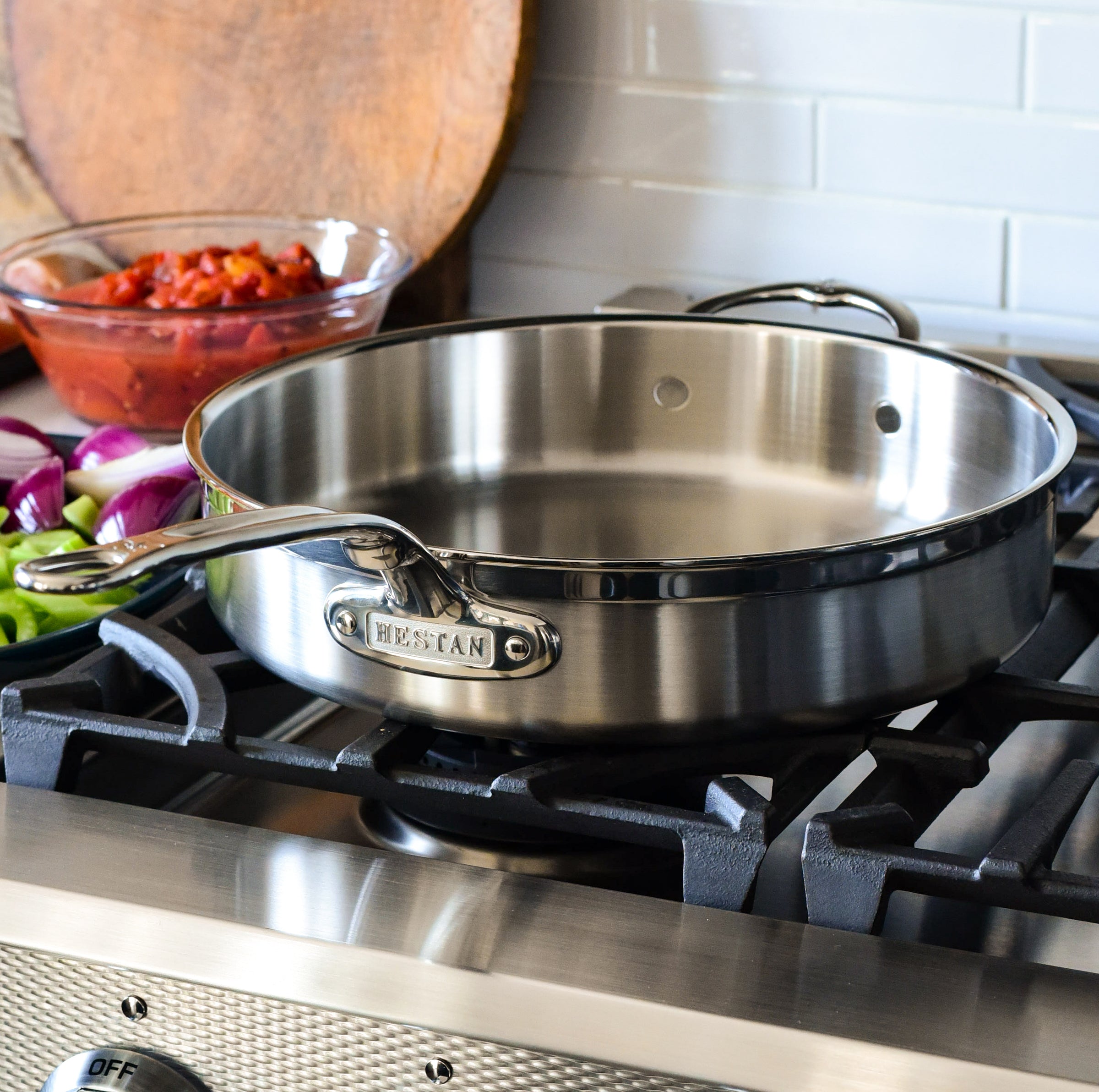 A stainless steel pan with handles is placed on a stovetop. In the background, there is a glass bowl of diced tomatoes and chopped purple onions on the counter.