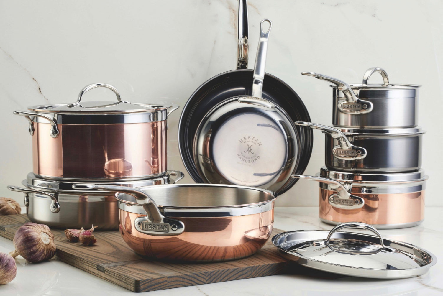A collection of copper and stainless steel pots and pans arranged on a wooden board. The set includes various sizes of saucepans and stockpots, each with lids. Three bulbs of garlic are placed on the board as decoration.