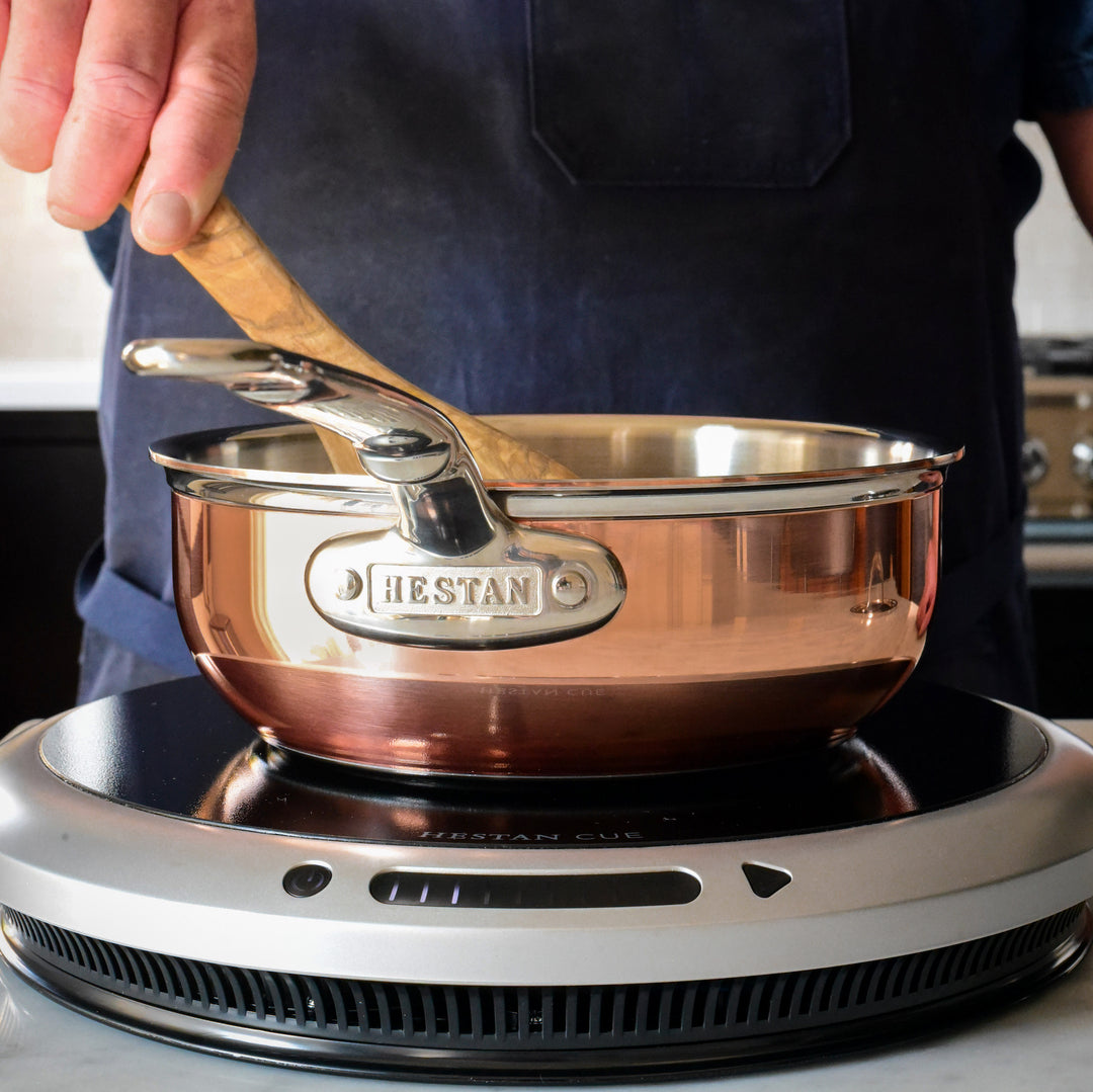A person wearing a dark blue apron stirs contents in a copper saucepan on an induction cooktop. The saucepan has the brand name HESTAN on its side. The background shows a blurred kitchen setting.
