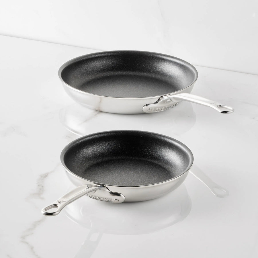 Two Thomas Keller Commercial Clad Stainless Steel TITUM® Nonstick Skillets rest on a white marble countertop, their handles pointing outward.