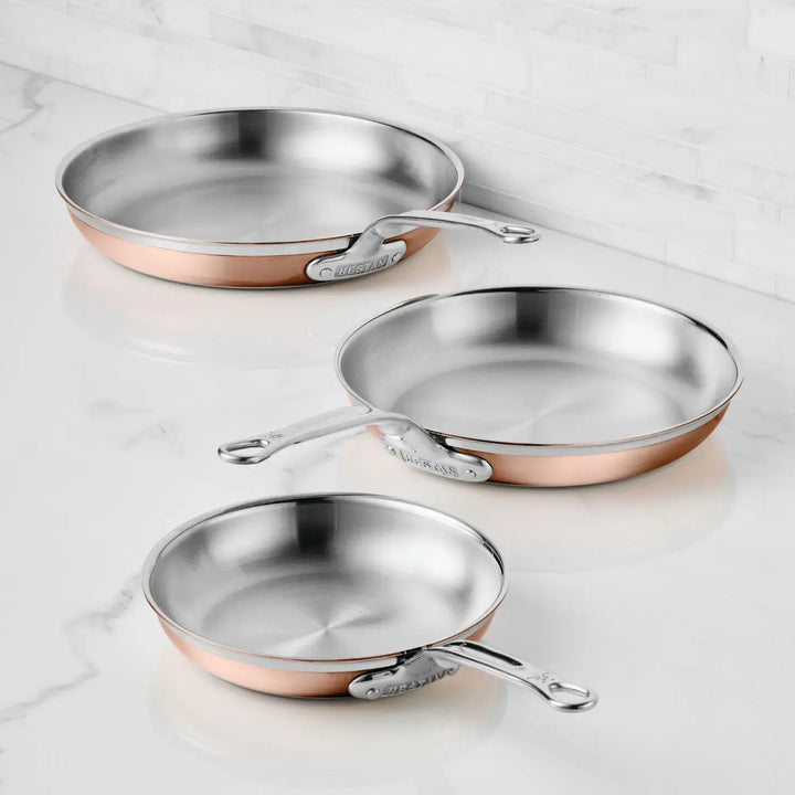 Three CopperBond Copper Induction Skillets in small, medium, and large sizes with stainless steel interiors and metal handles are arranged on a white marble countertop in front of a tiled wall.