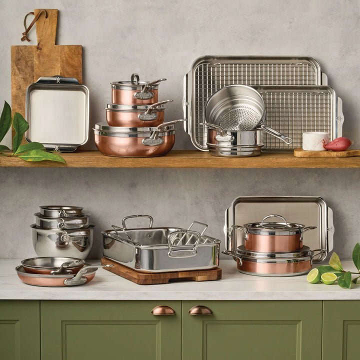 The CopperBond Copper Induction Set, 24-Piece, handcrafted in Italy, is stacked on a wooden shelf and green counter with baking trays, a colander, and utensils. Decorative plants and sliced limes add a fresh accent.