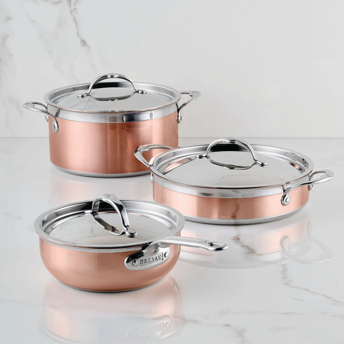 Three CopperBond Copper Induction Essential Set pots with stainless steel handles and lids are arranged on a marble countertop. This 6-piece set combines 100% copper core elegance with induction-ready performance.