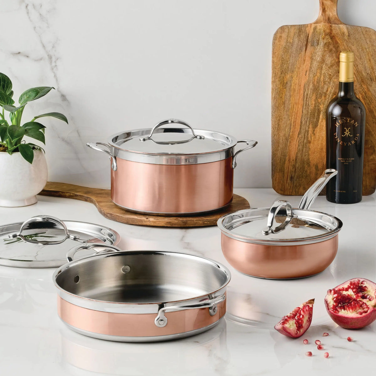 Three CopperBond Copper Induction Essential Set pieces with silver handles and lids are displayed on a white marble countertop beside a wooden cutting board, wine bottle, white potted plant, and cut pomegranate.