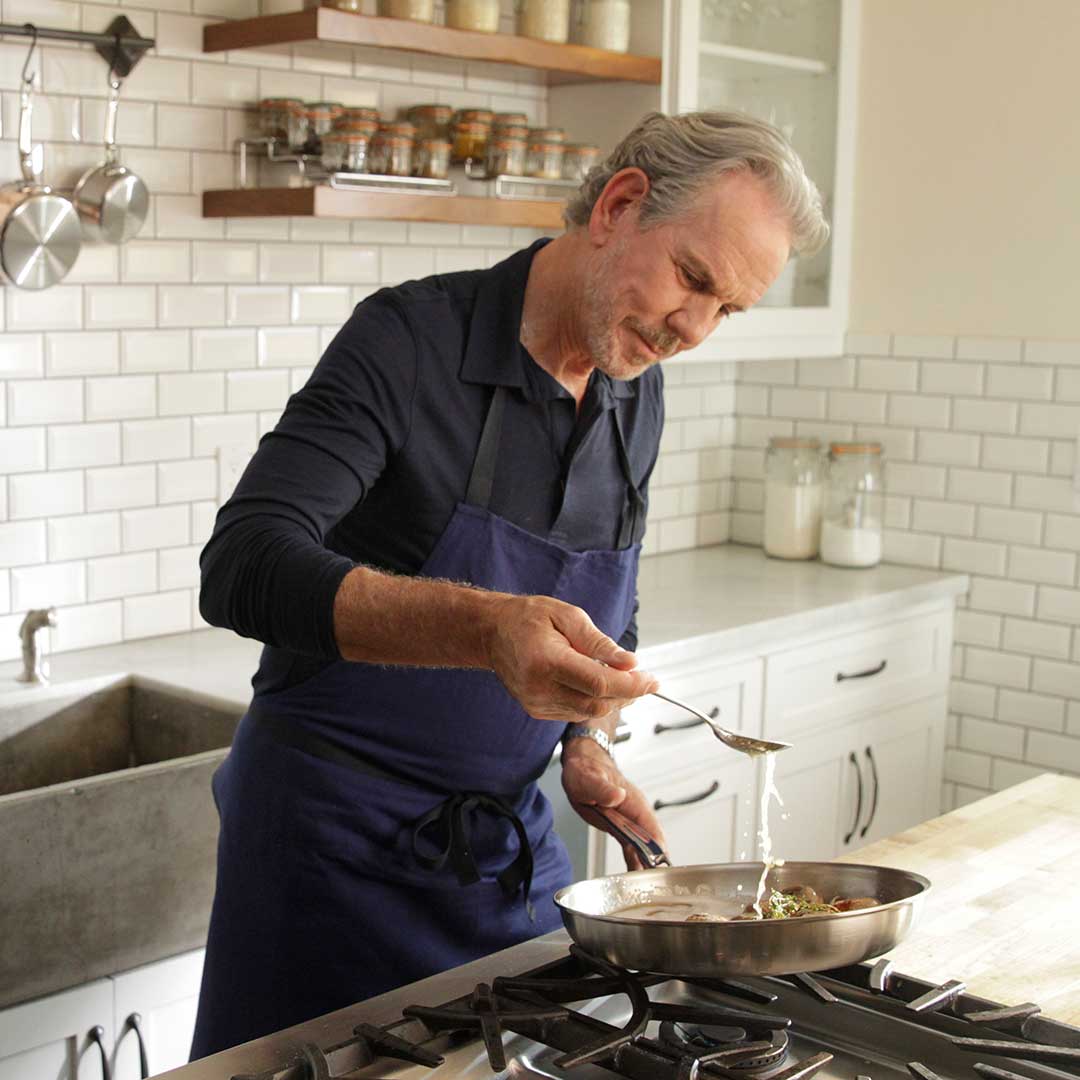A man in a navy apron is cooking in a kitchen. He is standing by a stove, using a spoon to add an ingredient to a frying pan. The kitchen has white subway tile walls, shelves with jars, and white cabinets.