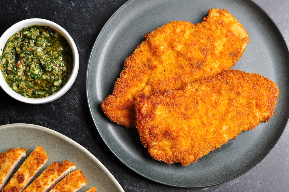 Breaded and fried chicken cutlets are served on a dark gray plate next to a small bowl of green herb sauce. Another plate with sliced cutlets is partially visible. The background is a dark surface.