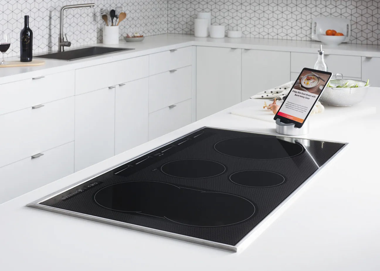 Modern kitchen with a sleek black induction cooktop on a white island countertop. A tablet with a recipe is propped up nearby. The background features white cabinets, a sink, and kitchen utensils against a geometric tile wall.
