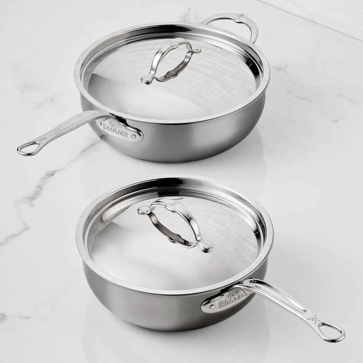 Two Titanium Essential Pans with lids by NanoBond sit on a white marble countertop. The stainless steel sauté pans feature long handles, matching metal lids with handles, and are marked with the NanoBond brand name.