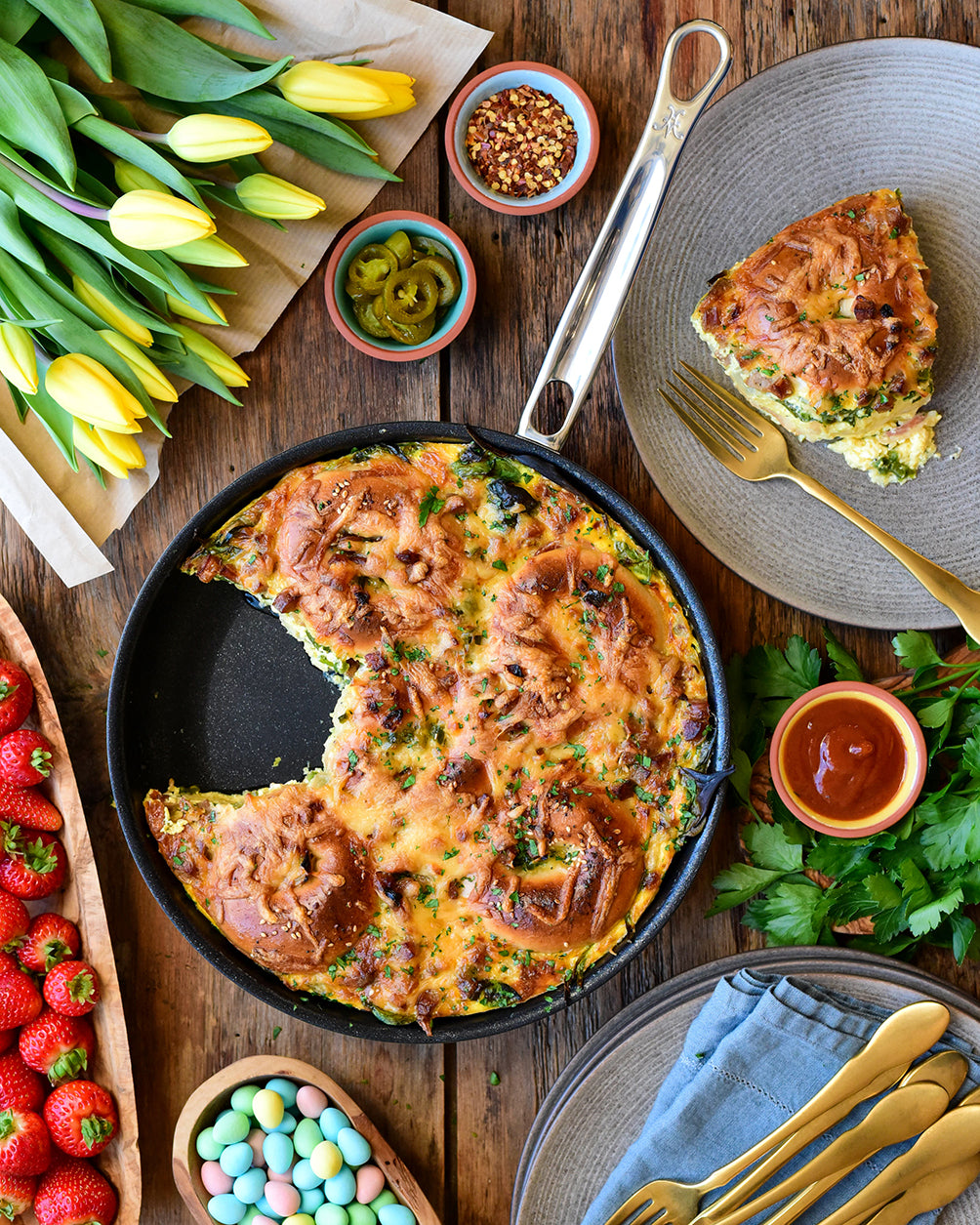A spinach and cheese quiche in a black skillet on a wooden table, with a slice served on a gray plate. Surrounding items include yellow tulips, strawberries, a bowl of mini chocolate eggs, jalapeños, chili flakes, a gold fork, and fresh parsley.