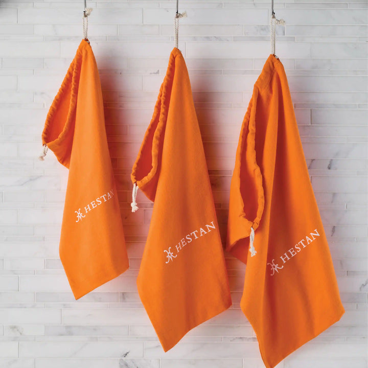 Three bright orange kitchen towels with HESTAN and a white logo hang on hooks against a white tiled wall, adding a pop of color beside your Accessories Flannel Cookware Storage Bags.