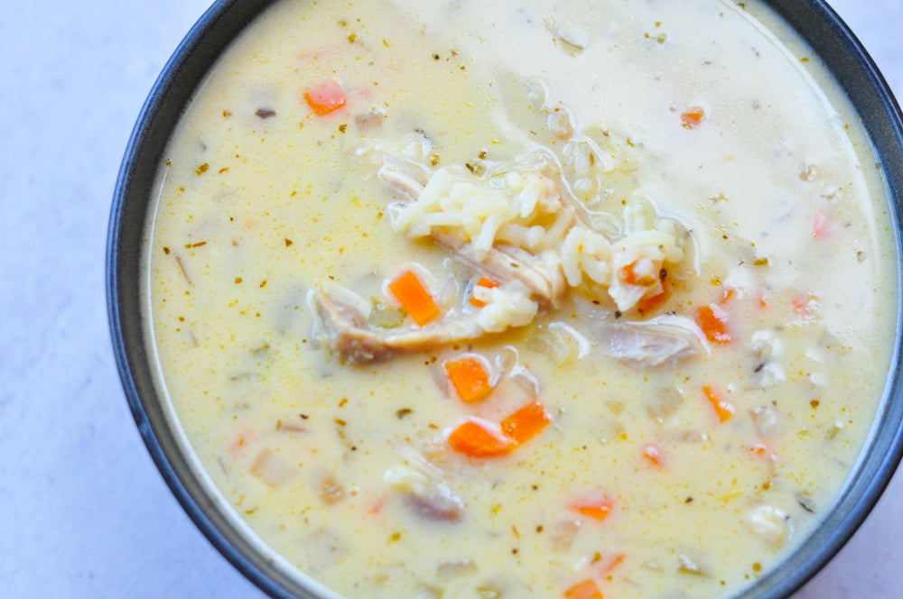 A bowl of creamy soup containing shredded chicken, carrots, rice, and herbs. The soup is light yellow with visible chunks of ingredients. The bowl is set against a plain, light-colored background.