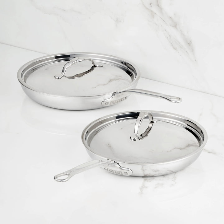Two ProBond® Luxe™ Polished Clad Stainless Steel Covered Skillets with matching lids and handles sit side by side on a white marble surface, gleaming in the light.