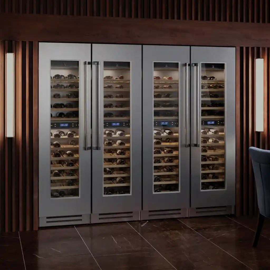 A modern wine cellar features four stainless steel wine refrigerators with glass doors, showcasing a collection of wine bottles. The room has wooden paneling, dim lighting, and a section of a dark chair is visible on the right.