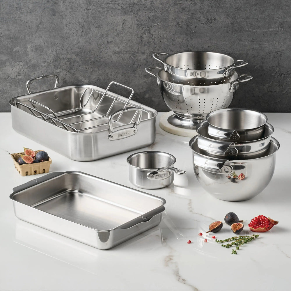 The Hestan Provisions Host and Roast 8-piece set, featuring stainless steel mixing bowls, colander, roasting and baking pans, and a measuring cup, is displayed neatly on a white counter with figs and herbs nearby.