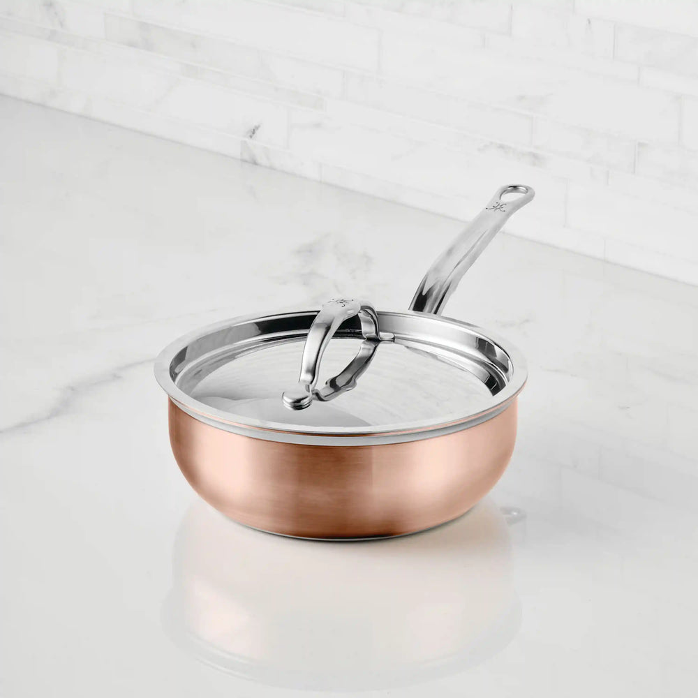 A CopperBond Copper Induction Saucier Pan, 2-Quart, with a stainless steel handle and lid sits on a white marble countertop, its sleek design highlighted against a backdrop of horizontally lined white marble.
