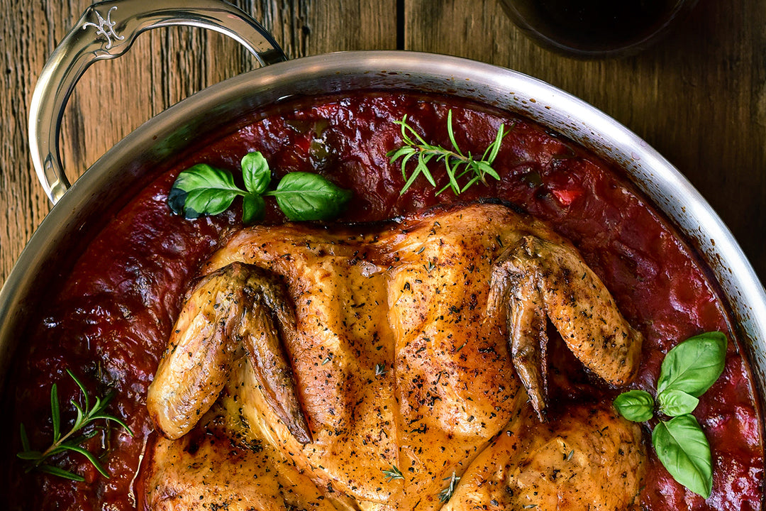 A roasted chicken seasoned with herbs is placed in a skillet filled with a tomato-based sauce. Fresh basil garnishes the dish. The skillet rests on a wooden surface.