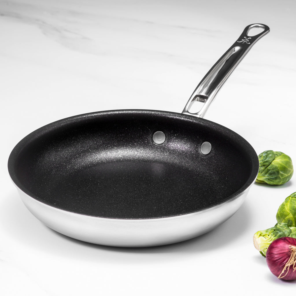 A stainless steel frying pan with a non-stick interior sits on a white marble surface. The handle has two rivets and a hanging hole. Brussels sprouts and a red onion are partially visible on the right side of the image.
