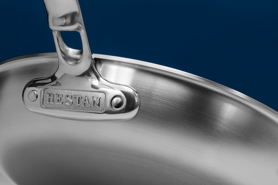 Close-up of a stainless steel frying pan on a blue background. The image shows a handle with a rectangular plate labeled HESTAN attached to the pan. The pans metallic surface is polished and reflective.