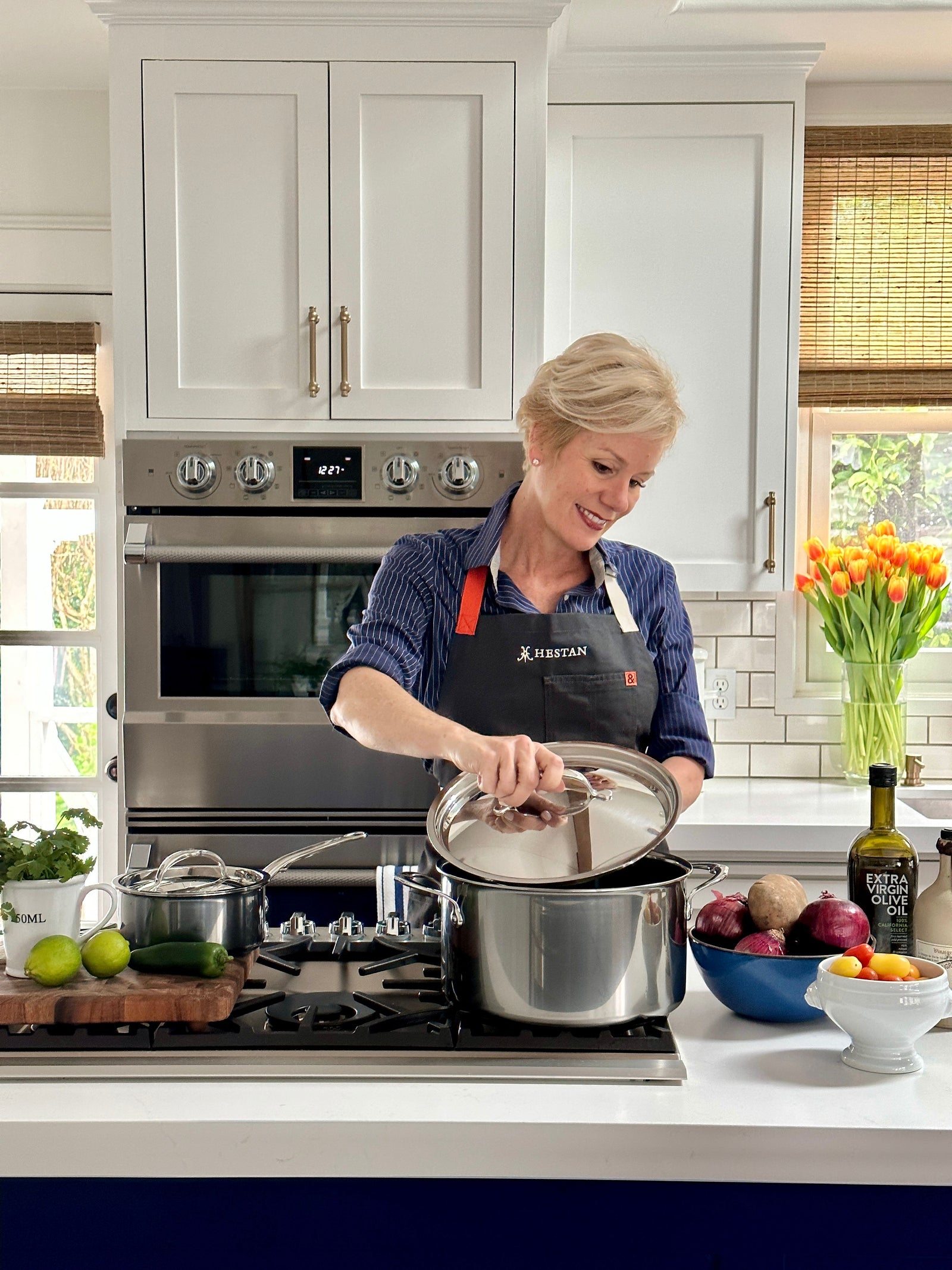 A person in a kitchen is standing by a stove, smiling while lifting the lid off a pot. The kitchen counter has various vegetables, a cutting board, and a bottle. A bouquet of tulips is placed near the window.