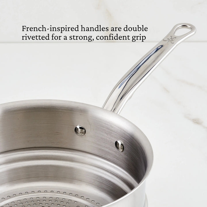 A saucepan from the Thomas Keller Commercial Clad Stainless Steel 29-Piece Cookware Set with double-riveted French-inspired handles sits on a white surface. Black text above reads, “French-inspired handles are double rivetted for a strong, confident grip.”.