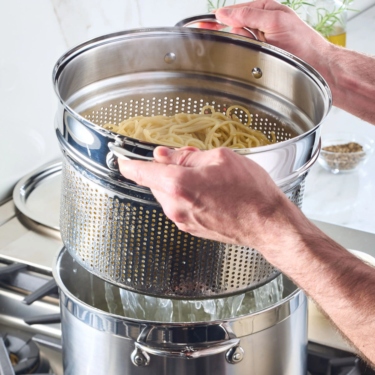 A person uses a Thomas Keller Commercial Clad Stainless Steel 29-Piece Cookware Set pasta strainer insert over a pot on the stove, draining cooked spaghetti as steam rises.