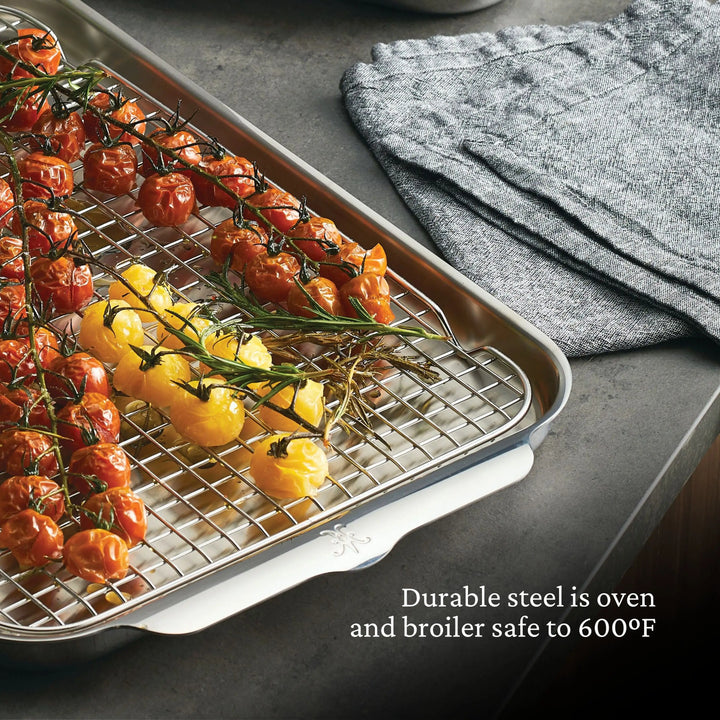 A Thomas Keller Commercial Clad 29-piece stainless steel baking tray holds roasted red and yellow tomatoes on the vine. The tray, set on a gray countertop by folded cloths, features durable steel that's oven and broiler safe to 600°F.