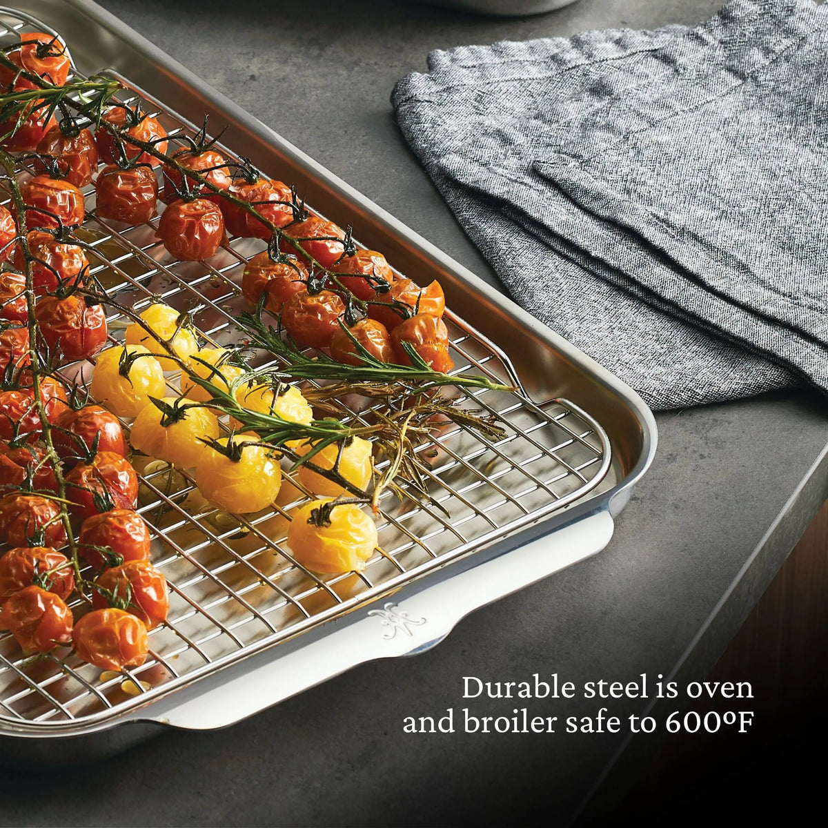 A Thomas Keller Commercial Clad 29-piece stainless steel baking tray holds roasted red and yellow tomatoes on the vine. The tray, set on a gray countertop by folded cloths, features durable steel that&