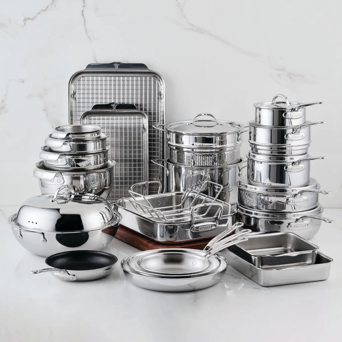The 29-piece Polished Clad Stainless Steel Pristine Set by ProBond® Luxe™ is displayed on a white surface, showcasing premium pots, pans, lids, casserole dishes, colander, roasting racks, and a cutting board against a marble backdrop.