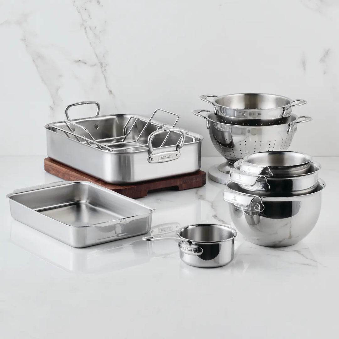 The Hestan Provisions Host and Roast 8-piece Set, featuring stainless steel cookware, mixing bowls, colanders, saucepans, a roasting pan, bakeware, and a wooden cutting board, is displayed on a white marble countertop.