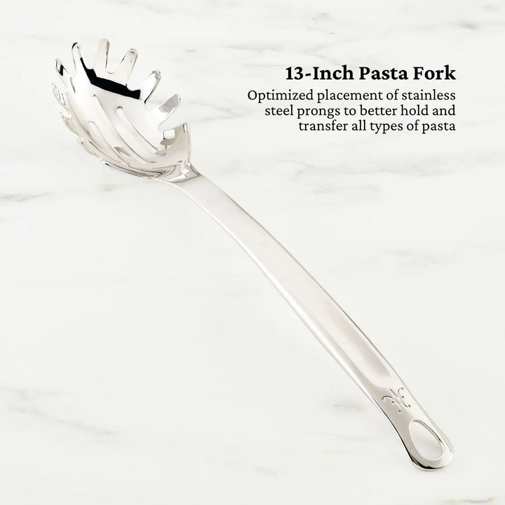 A Hestan Provisions pasta fork with pronged edges, designed for effortless pasta handling, sits on a light marble surface. The inscription beside it reads: 13-Inch Pasta Fork in tri-ply stainless steel. Its part of the OvenBond Epicurean 21-Piece Set.
