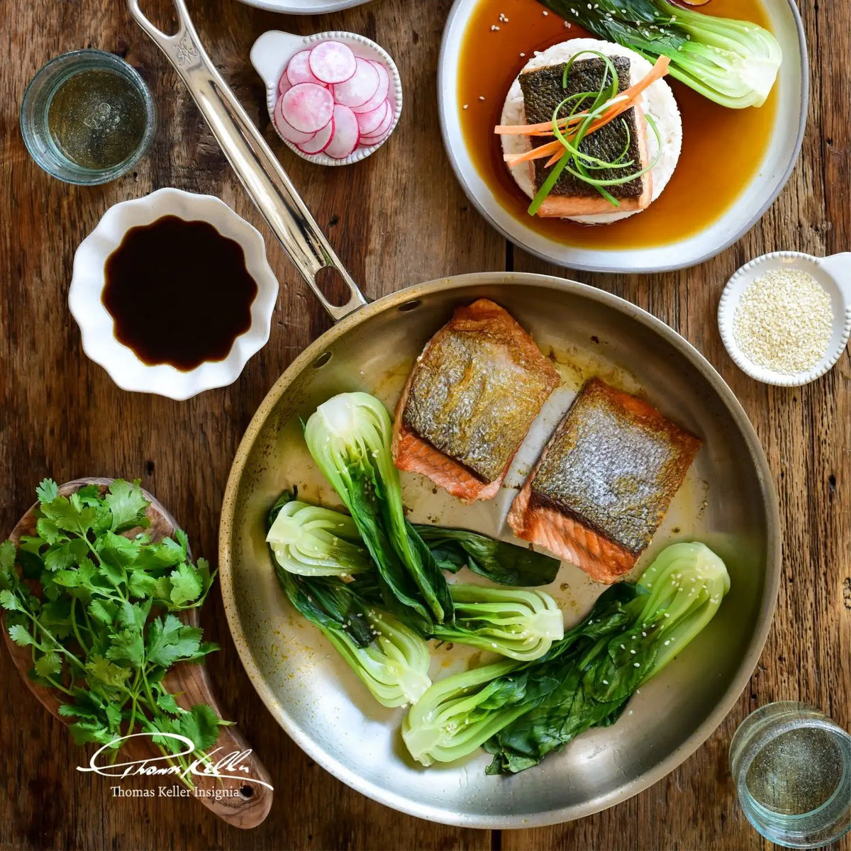 A meal of cooked salmon and bok choy in a pan from the Thomas Keller Commercial Clad Stainless Steel 10-Piece Cookware Set, served with fish fillet in broth, cilantro, radishes, soy sauce, sesame seeds, and water on a wooden table.