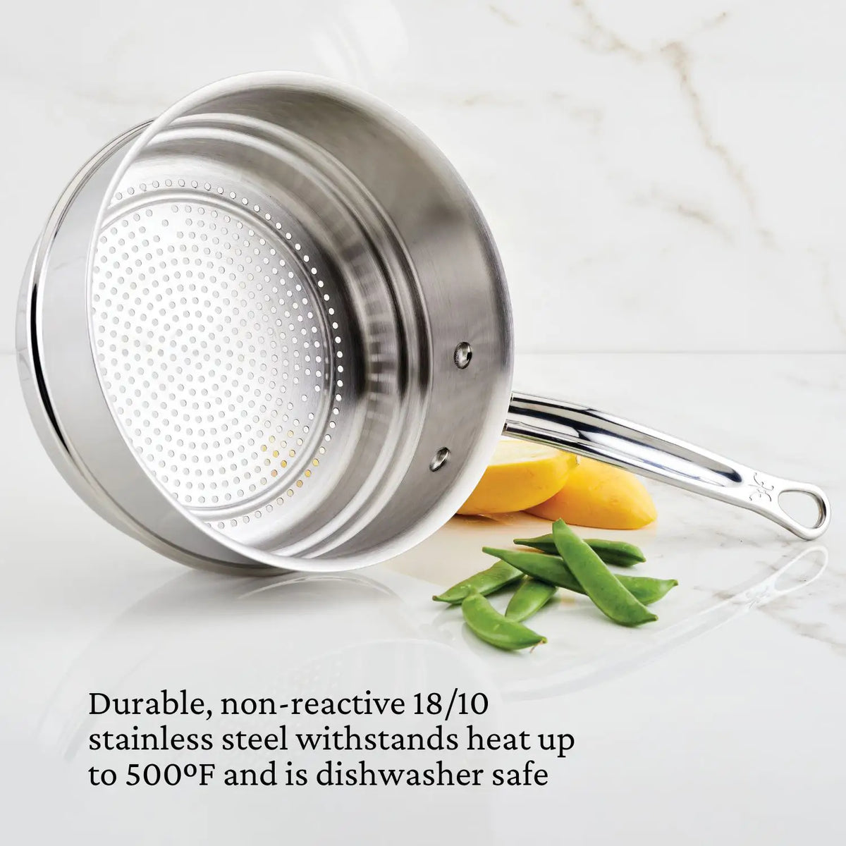 A Thomas Keller Commercial Clad Commercial Clad Stainless Steel 10-Piece Cookware Set saucepan with a perforated base rests on its side atop marble, with lemon wedges and green snap peas. Text notes its durable, heat-resistant, and dishwasher-safe design.