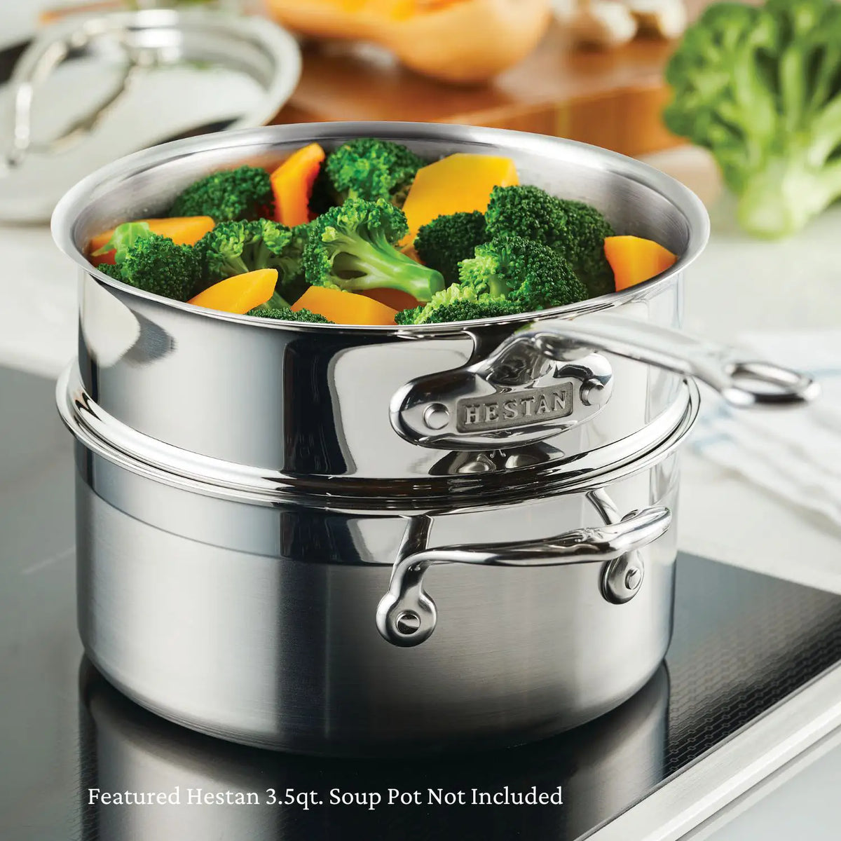 A stainless steel Thomas Keller Commercial Clad steamer pot from the Commercial Clad Stainless Steel 10-Piece Cookware Set sits on a stovetop with broccoli and carrots. Background features blurred veggies and kitchen items. Soup pot not included.